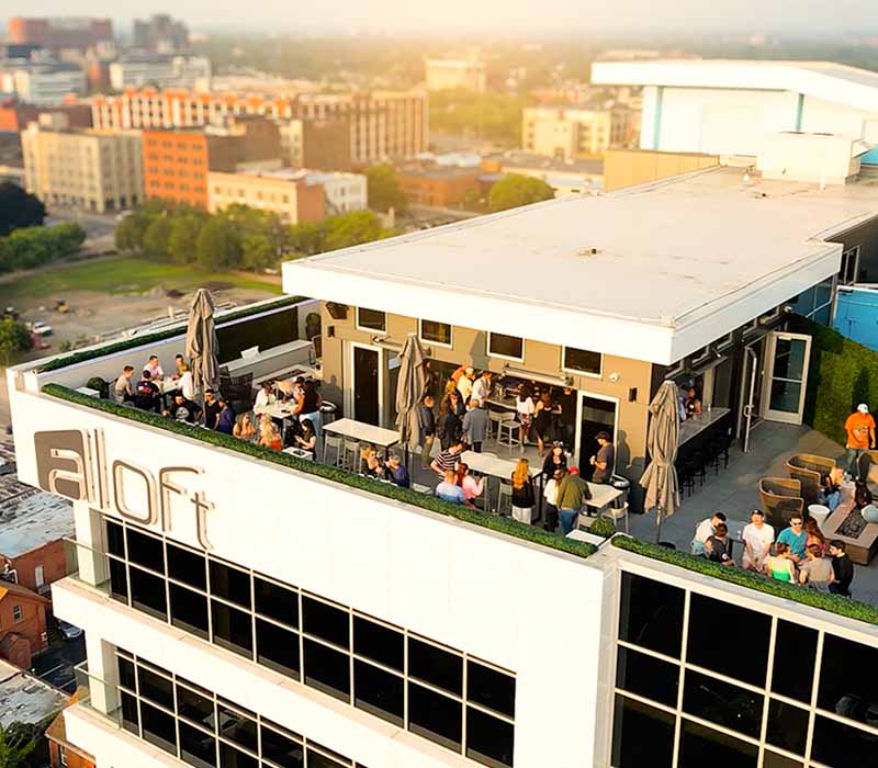 View of the 500 Pearl building rooftop, featuring Patrick’s Rooftop Bar with guests enjoying cocktails near glass railings. The large illuminated Aloft hotel sign is prominently displayed on the building, while the Buffalo city skyline stretches across the background under a vibrant sky, highlighting the lively downtown atmosphere.